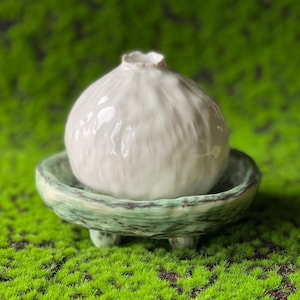 以下が含まれることがあります： テクスチャ加工された表面を持つ白いセラミックの花瓶が、3本の脚が付いた緑色のセラミックの台座の上に置かれています。花瓶と台座は緑色の苔の上に置かれています。