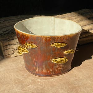 Op de afbeelding: Een keramische plantenbak met een bruine houtnerftextuur en verschillende geelgouden paddenstoelvormige accenten. De binnenkant is lichtcrème van kleur. De plantenbak staat op een houten oppervlak.