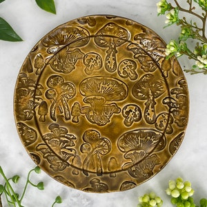 May include: A brown ceramic plate with a raised design of mushrooms in various shapes and sizes. The plate has a glossy finish and is perfect for serving snacks or appetizers.