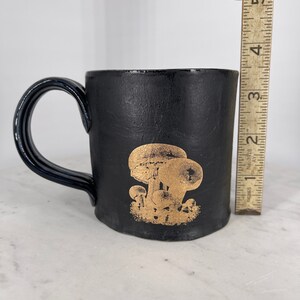 May include: Black ceramic mug with a gold mushroom design. The mug has a rounded handle.