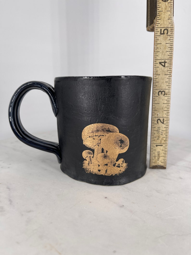 May include: Black ceramic mug with a gold mushroom design. The mug has a rounded handle.