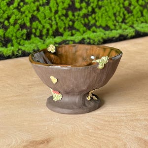 Könnte beinhalten: Eine kleine, braune Keramikschale mit einem Sockel. Die Schale hat eine strukturierte Oberfläche und ist mit kleinen, grünen und gelben Akzenten verziert.