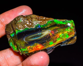 Puede incluir: Una gema de ópalo en bruto con colores vibrantes de verde, naranja y azul. El ópalo se sostiene en una mano, mostrando su juego de colores iridiscente. La piedra tiene una matriz oscura y natural.