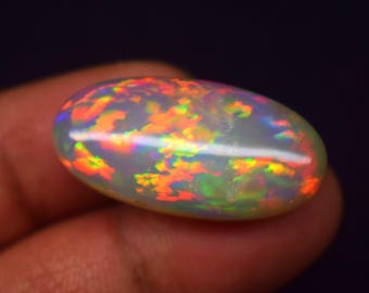 May include: A polished, oval-shaped opal gemstone. The opal displays a milky white base color with vibrant flashes of orange, red, yellow, and green, creating a play-of-color effect. The gemstone is held in a hand against a dark background.