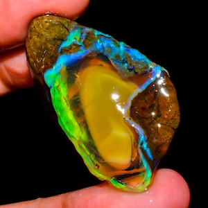 Puede incluir: Una vibrante piedra preciosa de ópalo en bruto con una mezcla de colores. La piedra presenta un núcleo central amarillo, rodeado por una capa translúcida con destellos verdes y azules. La capa exterior es de color marrón oscuro con una textura rugosa.