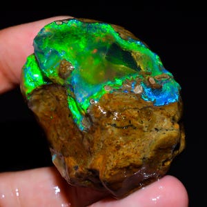 May include: A raw, unpolished boulder opal specimen featuring a prominent, vibrant play-of-color with intense neon green and electric blue flashes set against a textured, earthy brown ironstone matrix with natural crevices and irregular surface contours.
