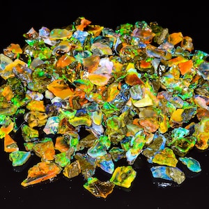 May include: A large pile of colorful opal chips, with shades of green, blue, orange, and yellow. The chips are iridescent and have a variety of shapes and sizes.