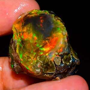 Könnte beinhalten: Ein roher, ungeschliffener Opal-Edelstein mit einem lebendigen Farbspiel. Der Opal zeigt Blitze von Orange, Grün und Gelb auf einem dunklen, fast schwarzen Untergrund. Der Stein wird in einer Hand gehalten und zeigt seine natürliche, rohe Textur.