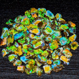 May include: A pile of rough, unpolished opal gemstones. The opals are a mix of green, blue, yellow, and orange colors. The opals are iridescent and have a variety of shapes and sizes.