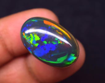 Puede incluir: Una piedra preciosa de ópalo negro pulido de forma ovalada. El ópalo muestra destellos de color vibrantes, incluyendo azul, verde, naranja y amarillo, sobre un fondo oscuro. La gema se sostiene entre los dedos, mostrando sus cualidades iridiscentes.