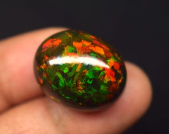Puede incluir: Una piedra preciosa pulida, de forma ovalada, con un color de base oscuro. La piedra muestra vibrantes colores internos de rojo, naranja y verde, creando un patrón de mosaico. La gema se sostiene sobre un fondo oscuro.