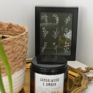 May include: A framed print with a green background and white line drawings of various flowers and leaves. The frame is black and the print is sitting on a white surface next to a black candle jar with a gold lid and a gold tray.