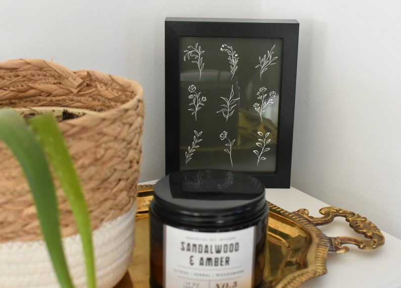 May include: A framed print with a green background and white line drawings of various flowers and leaves. The frame is black and the print is sitting on a white surface next to a black candle jar with a gold lid and a gold tray.