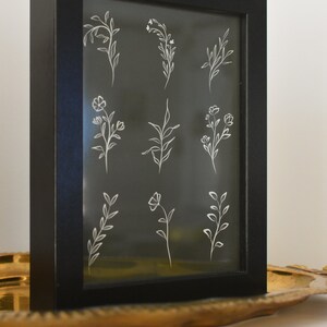 May include: A black framed picture with a white background and nine white line drawings of different flowers and plants.