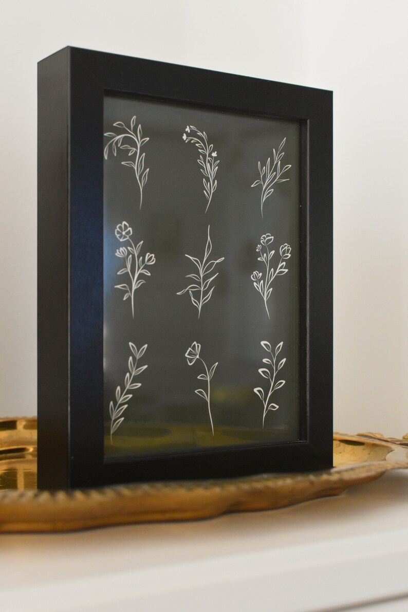 May include: A black framed picture with a white background and nine white line drawings of different flowers and plants.