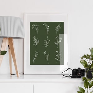 May include: A white framed print with a dark green background and white line drawings of various floral designs. The print is sitting on a white shelf with a lamp and a potted plant on either side.