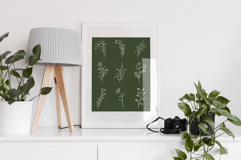 May include: A white framed print with a dark green background and white line drawings of various floral designs. The print is sitting on a white shelf with a lamp and a potted plant on either side.