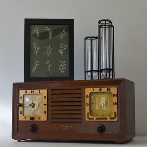 May include: A vintage wooden radio with two clock faces and a black framed print of white floral illustrations above it. The radio has a brown wooden case with a metal grille and two knobs on the front. The clock faces are white with black numbers and hands. The print is on a dark green background.
