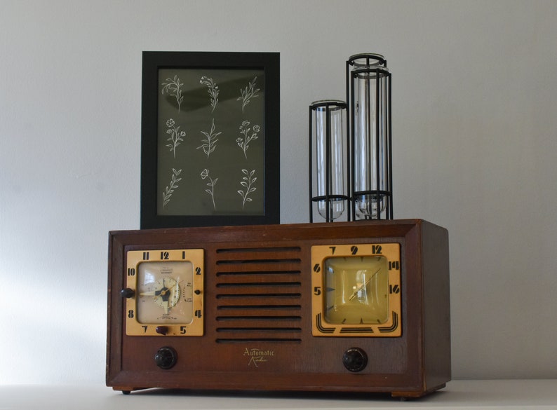 May include: A vintage wooden radio with two clock faces and a black framed print of white floral illustrations above it. The radio has a brown wooden case with a metal grille and two knobs on the front. The clock faces are white with black numbers and hands. The print is on a dark green background.