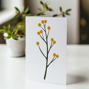 Op de afbeelding: Een witte wenskaart met een minimalistisch botanisch ontwerp. De illustratie toont een donkergroene stengel met verschillende geel-oranje cirkelvormige knoppen. De kaart staat tegen een vage achtergrond met een kleine plant in een pot.