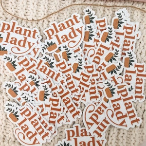 May include: A pile of white stickers with the text "plant lady" in brown lettering. The stickers feature a small potted plant with green leaves and a brown pot.