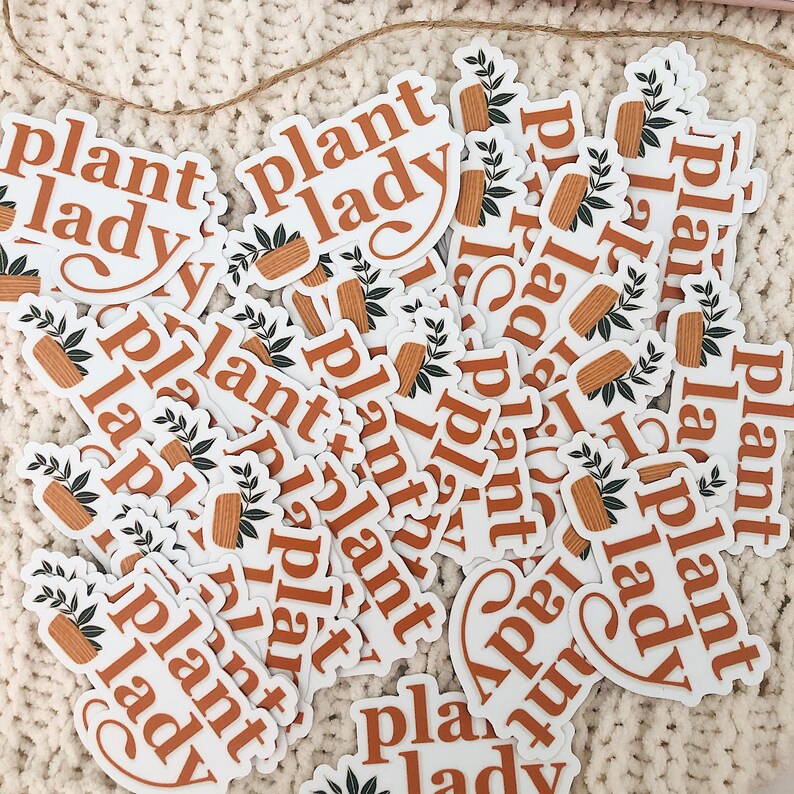 May include: A pile of white stickers with the text "plant lady" in brown lettering. The stickers feature a small potted plant with green leaves and a brown pot.