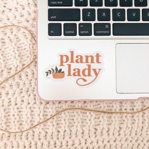 May include: A white laptop with a sticker that says "plant lady" in orange text with a small potted plant illustration. The laptop is on a white fuzzy surface.