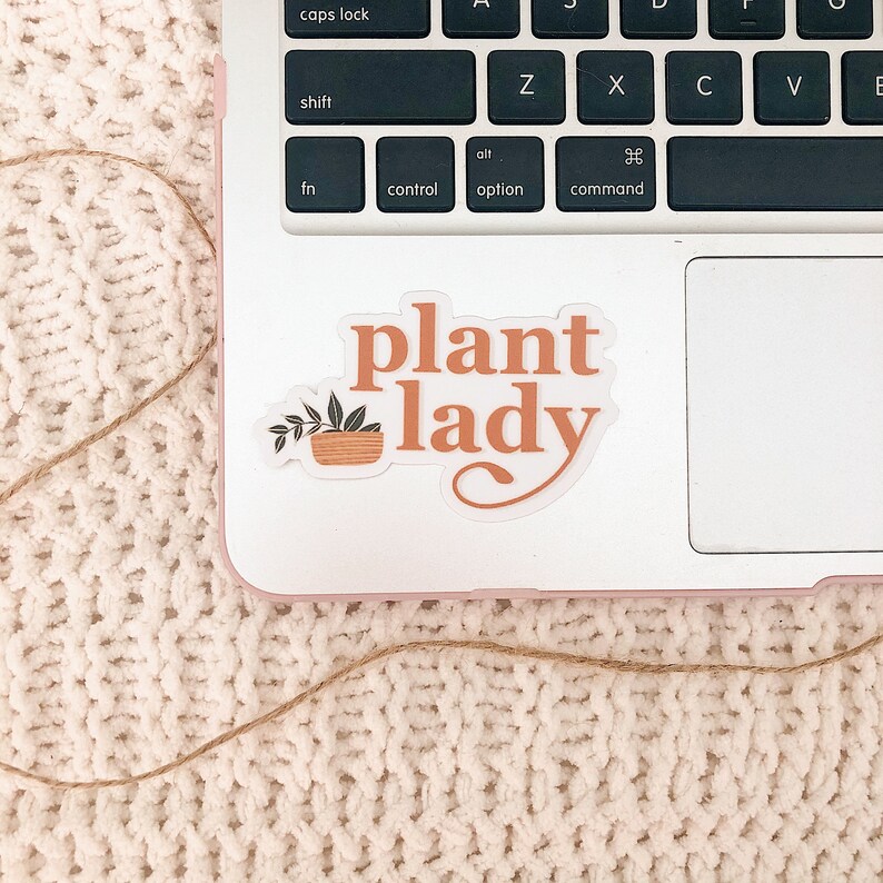 May include: A white laptop with a sticker that says "plant lady" in orange text with a small potted plant illustration. The laptop is on a white fuzzy surface.