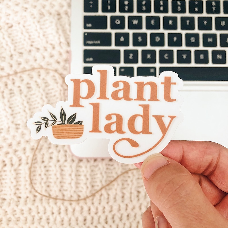 May include: A white sticker with the text "plant lady" in orange lettering. A small potted plant with green leaves is to the left of the text.