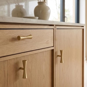 Può includere: Mobili in legno di rovere chiaro dotati di tre maniglie per armadietti in metallo color ottone. La ferramenta presenta una distinta forma a clessidra con un centro cilindrico liscio ed estremità svasate, montata orizzontalmente sul cassetto e verticalmente sulle ante.