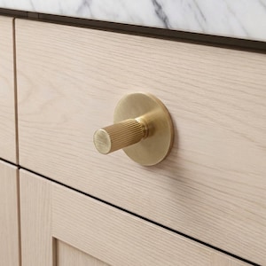 Könnte beinhalten: Nahaufnahme eines goldfarbenen Schrankknaufs mit einem zylindrischen, geriffelten Griff und einer kreisförmigen Basis. Der Knauf ist an einer hellen Holzschranktür befestigt. Die Schranktür ist Teil eines größeren Schranks.