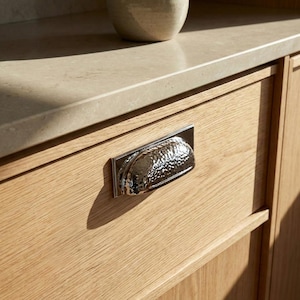 May include: A close-up of a wooden cabinet drawer with a silver, hammered-texture handle. The handle is rectangular with a curved, semi-circular grip. The wood grain is visible, and the cabinet is light-colored.