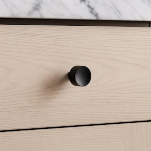 May include: A black, cylindrical cabinet knob is mounted on a light-colored wooden drawer. The knob has a matte finish and a simple, modern design. The drawer is part of a larger piece of furniture.