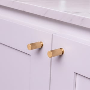 May include: Two gold cabinet knobs with a ribbed design are mounted on white cabinet doors.