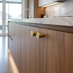 May include: Close-up of a kitchen cabinet with two brass-colored, cylindrical cabinet pulls. The cabinet doors are made of wood with a natural grain. A marble countertop is visible in the background.