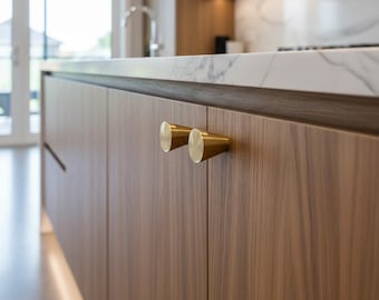 Puede incluir: Primer plano de un mueble de cocina con dos tiradores cilíndricos de color latón. Las puertas del armario son de madera con veta natural. En el fondo se ve una encimera de mármol.