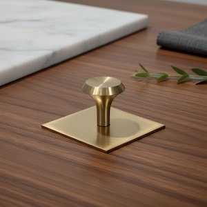 May include: A contemporary brass cabinet knob with a square base. The knob features a rounded top and a flared stem. The hardware is set against a dark wood surface, with a marble slab and a grey cloth in the background.