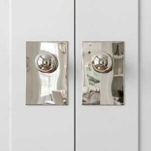 May include: Close-up of two cabinet doors with polished silver hardware. Each door has a rectangular backplate with a rounded, spherical knob. The hardware reflects the surroundings, creating a bright and modern aesthetic.