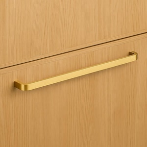 May include: A close-up of a wooden cabinet with a gold-colored rectangular handle. The handle is mounted horizontally and has a smooth, modern design. The cabinet has a light brown wood grain finish.