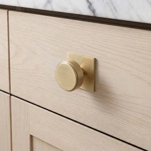 Op de afbeelding: Een close-up van een goudkleurige kastknop met een vierkante achterplaat en een ronde knop. De knop is gemonteerd op een lichtgekleurde houten kastdeur. De kast maakt deel uit van een groter meubelstuk.