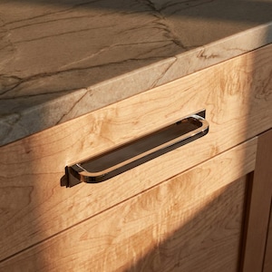 Könnte beinhalten: Nahaufnahme einer Holzschublade mit einem rechteckigen, schwarz-goldfarbenen Griff. Die Schublade ist aus hellem Holz gefertigt, und die Arbeitsplatte darüber ist hellbraun mit dunkleren Maserungen.