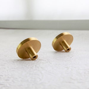 May include: Two gold-toned round cabinet knobs with a smooth, polished finish. The knobs have a cylindrical stem and a threaded screw hole on the bottom.