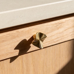 May include: A gold-toned metal cabinet knob with a faceted, hourglass-like shape featuring a central point and radiating ridges. The knob is mounted on a light-coloured wooden drawer front with a visible grain pattern and a smooth, light-coloured countertop.
