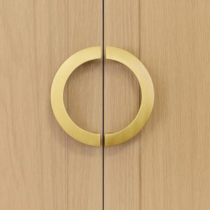 May include: Close-up of a light wood cabinet door with two halves of a gold-coloured, circular handle. The handle forms a complete circle when the doors are closed. The wood grain is visible.