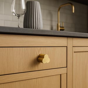 Könnte beinhalten: Nahaufnahme eines Küchenschranks aus Holz mit einem goldfarbenen, kleeblattförmigen Schubladengriff. Der Schrank hat eine helle Holzmaserung. Ein Weinglas, eine graue Vase und ein goldener Wasserhahn sind im Hintergrund zu sehen.