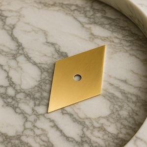 以下が含まれることがあります： 単一の円形の穴がある、金色のひし形をしたキャビネットプル。 金具は、大理石模様の表面に配置されており、テクスチャと色のコントラストを生み出しています。 プルは、引き出しまたはキャビネット用である可能性があります。