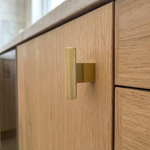 May include: A gold-toned T-bar cabinet knob with a cylindrical, knurled texture on the handle and a smooth, rectangular base plate. The hardware is mounted onto a light-colored, vertical-grained wooden cabinet door surface.