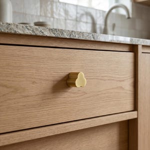 May include: Close-up of a wooden drawer with a gold-colored, three-lobed cabinet knob. The drawer is made of light-colored wood with visible grain. The knob is centered on the drawer, providing a simple, yet elegant design element. The background includes a countertop and a faucet.