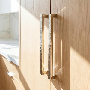Könnte beinhalten: Nahaufnahme von zwei goldfarbenen Schrankgriffen mit gehammerter Textur, montiert an hellen Holzschranktüren. Die Griffe sind rechteckig und vertikal ausgerichtet. Natürliches Licht wirft Schatten auf das Holz.