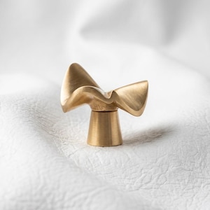May include: A gold-toned metal cabinet knob featuring a sculptural, wave-like top with a brushed finish. The hardware has a flared, cylindrical base that supports the wide, curved upper section, creating a distinct, fluid form for furniture.